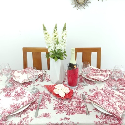 Mesa de jantar posta com toalha, guardanapos e almofadas vermelhas e brancas, pratos, copos, talheres, garrafa de champanhe no balde e vaso com flores brancas.