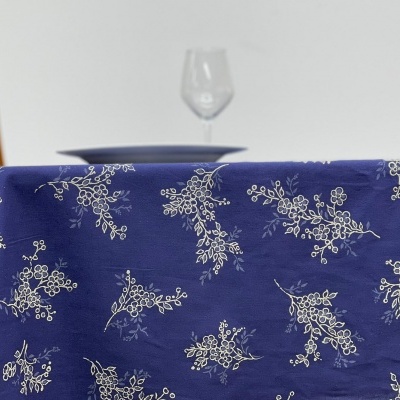 Toalha de mesa azul com padrão floral branco sobre mesa com peça de louça e copo