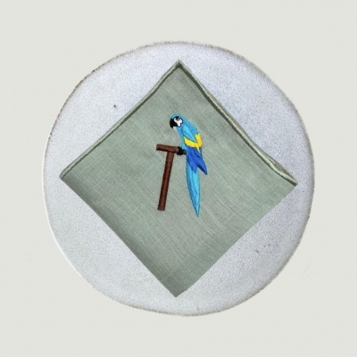 Guardanapo verde-oliva bordado com papagaio azul numa base redonda branca.