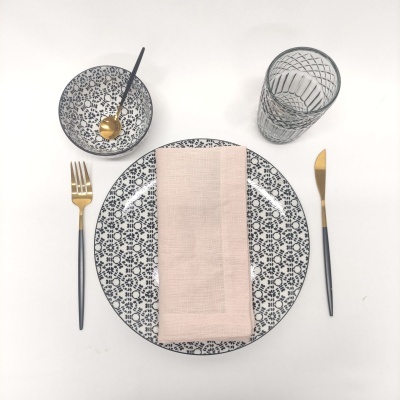 Conjunto de louça com padrão geométrico preto e branco, guardanapo rosa, talheres dourados e copo de vidro