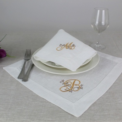 Guardanapo e toalha branca bordados com letras douradas com talheres, copo e flores roxas