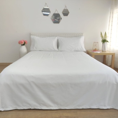 Cama com lençol e fronhas brancas e cabeceira branca trançada numa sala decorada com espelhos geométricos e mesa de cabeceira de madeira