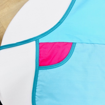 Avental azul claro com bolso rosa e bordas azul em tecido sobre fundo branco e madeira