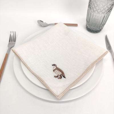 Jogo de jantar com prato branco, guardanapo bordado com ave, talheres com cabo de madeira e copo alto transparente.