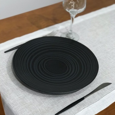 Prato negro com padrão circular, faca preta e copo de vinho em toalha branca sobre mesa de madeira