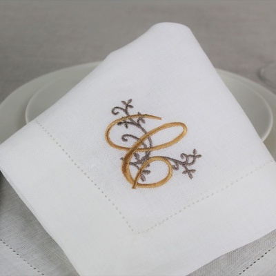 Guardanapo branco com bordado castanho e dourado num prato branco com talheres ao lado e copo de vidro no fundo