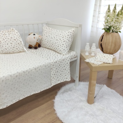 Conjunto de roupa de cama infantil branca com padrão de folhas pequenas sobre cama branca ao lado de mesa de madeira clara com vaso e flores brancas