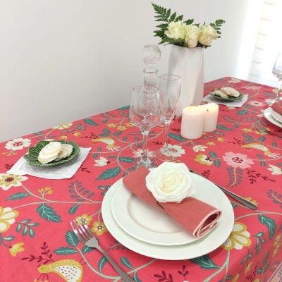 Mesa posta com toalha floral vermelha, pratos brancos, guardanapo coral com flor branca, copos de vidro, velas, jarro com flores brancas