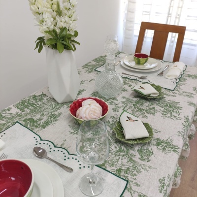 Mesa de jantar decorada com toalha verde e branca, pratos brancos com interior vermelho, copos de vidro, guardanapos bordados e vaso de flores brancas