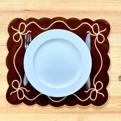 Prato branco com talheres sobre individual de tecido castanho com bordados bege em mesa de madeira clara