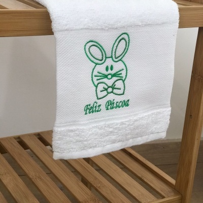 Toalha branca com bordado verde de coelho e texto Feliz Páscoa pendurada em prateleira de madeira