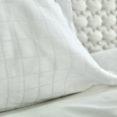 Almofada branca com padrão de quadrícula em tecido, sobre superfície branca com manta texturada ao fundo.