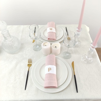 Mesa posta com pratos brancos, guardanapos cor de rosa, talheres pretos e dourados, velas e copos de vidro