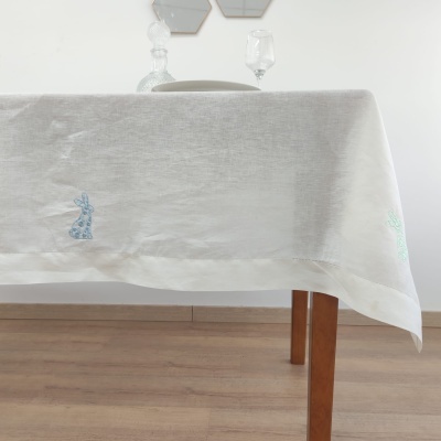 Toalha de mesa branca com bordados azuis e verdes em mesa de madeira com garrafa e copo