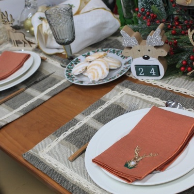 Mesa de jantar decorada para Natal com pratos brancos, guardanapos cor de ferrugem, talheres dourados, e ornamentos natalícios em madeira