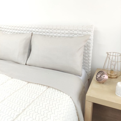 Cama com cabeceira branca entrelaçada, roupa de cama cinza e branca, mesa de cabeceira com relógio cor de rosa, vela e lâmpada metálica