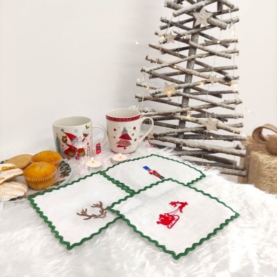 Canecas natalícias, bolos, velas e jogos americanos bordados sobre superfície branca com árvore de Natal de ramos ao fundo