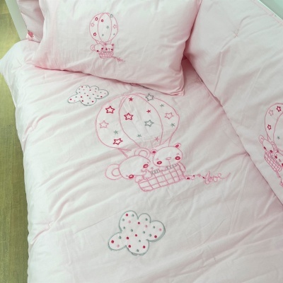 Lençóis de cama rosa claro com desenho bordado de balão de ar quente com dois ursos e nuvens no tecido.
