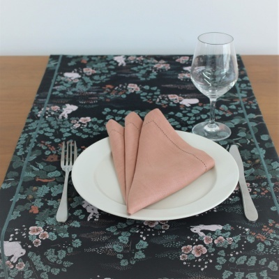 Conjunto de jantar com prato branco, guardanapo rosa, talheres prateados e copo de vidro sobre toalha de mesa preta com padrão floral.