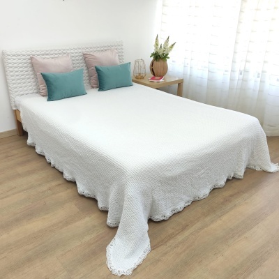 Cama com colcha branca de crochê e almofadas coloridas num quarto com chão de madeira e mesa lateral