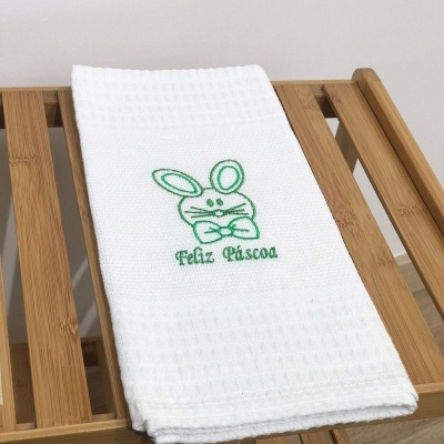 Toalha branca bordada com coelho e texto 'Feliz Páscoa' sobre suporte de madeira