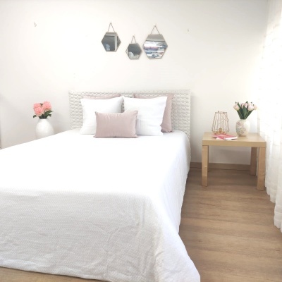 Cama com lençol branco e almofadas, mesas de cabeceira com vasos de flores, espelhos hexagonais na parede branca