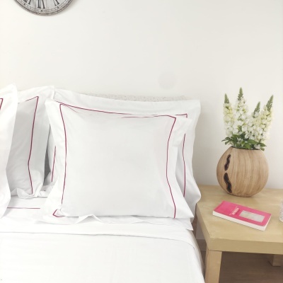 Almofadas brancas com bordas vermelhas no quarto ao lado de vaso de madeira com flores brancas e agenda rosa.