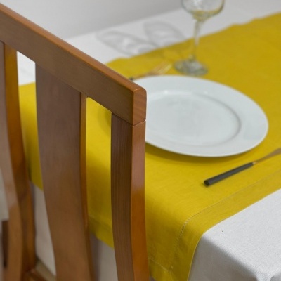 Mesa posta com toalha branca e toalha amarela, prato branco, faca preta, copo de vinho e cadeira de madeira.