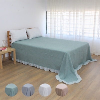 Cama com lençol azul e saia branca em quarto com mesa de madeira e cortinas brancas