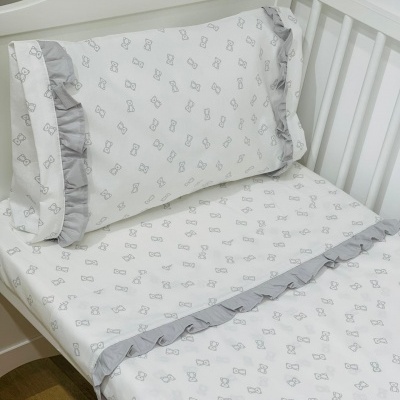 Lençol e fronha de algodão branco com padrão de laços cinzentos e folhos cinzentos numa cama de bebé branca