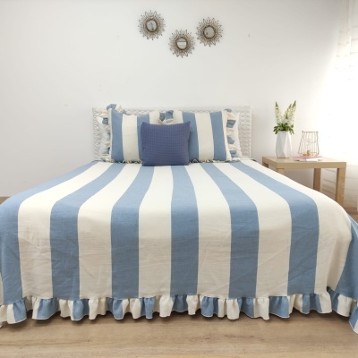 Cama com colcha às riscas azul claro e branco e almofadas, mesa lateral com vaso e parede branca com espelhos redondos.