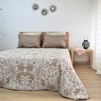 Quarto com cama de madeira, roupa de cama floral bege e castanha, almofadas castanhas, mesa lateral com objeto preto, bata branca pendurada, cortinas brancas e decoração de parede metálica