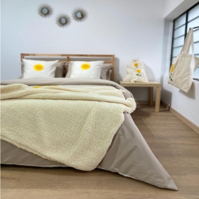 Quarto com cama de roupa de cama cinza e edredão creme, almofadas com girassol, mesa e bolsa pendurada.