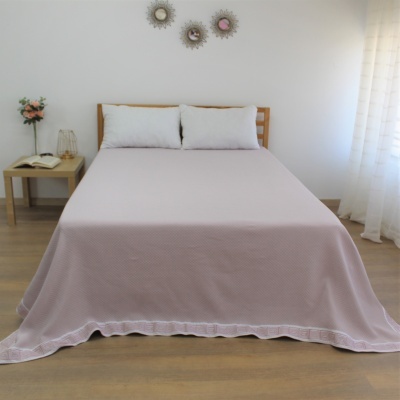Cama casal com cobre-leito rosa claro, almofadas brancas, mesa cabeceira com flores e candeeiro, parede branca decorada.