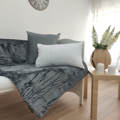 Cobertor cinza felpudo sobre cama com almofadas, mesa bege com vaso de flores brancas e relógio na parede