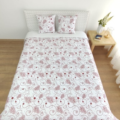 Cobertura de cama branca com padrão floral vermelho e rosa, duas almofadas, cama com cabeceira branca, mesa de madeira com vaso e livros