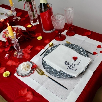 Mesa posta com toalha vermelha, prato cinza, guardanapo branco com desenho, talheres dourados e decoração com velas e pétalas vermelhas.