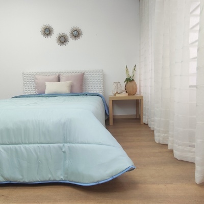 Cama com edredão azul claro e almofadas, mesa de cabeceira com jarro e flores, cortinas brancas, parede e chão claro