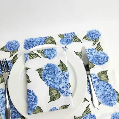 Guardanapo e toalha com padrão de flores hortênsia azuis em fundo branco em conjunto com prato e talheres.