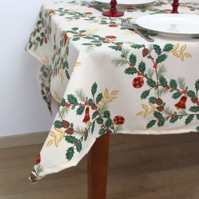 Toalha de mesa creme com padrão natalício de folhas, pinhas, sinos e bolas vermelhas sobre mesa de madeira.