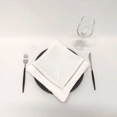 Mesa posta com prato preto, guardanapo branco bordado, talheres de cabo preto e copo de vidro.