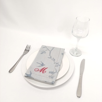Conjunto de jantar com prato branco, talheres metálicos, copo de vidro e guardanapo cinzento com padrão floral e letra 'M'.