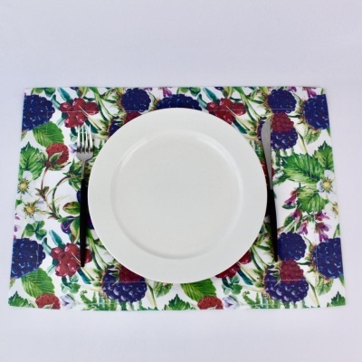 Conjunto debaixo pratos com padrão de frutos vermelhos e folhas verdes, prato branco redondo e talheres prateados