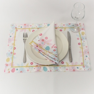 Jogo americano e guardanapo com padrão floral, prato branco, talheres e copo de vidro sobre mesa branca.