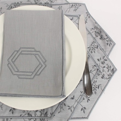 Mesa posta com prato branco, guardanapo cinzento bordado, faca metálica e base de prato com padrão floral cinzento