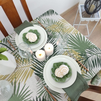 Mesa posta com toalha de folhas verdes, pratos brancos, guardanapos verdes, velas acesas e talheres castanhos.
