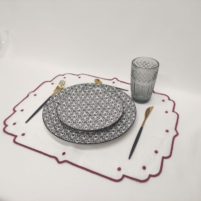 Conjunto de jantar com pratos decorados, talheres com cabos pretos e dourados, copo cinzento e base de tecido branca com bordados vermelhos