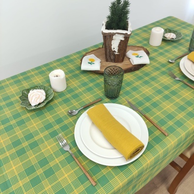 Mesa posta com toalha verde e amarela, pratos, talheres, guardanapo amarelo, copo verde, velas brancas e decoração com tronco e almofadas bordadas.