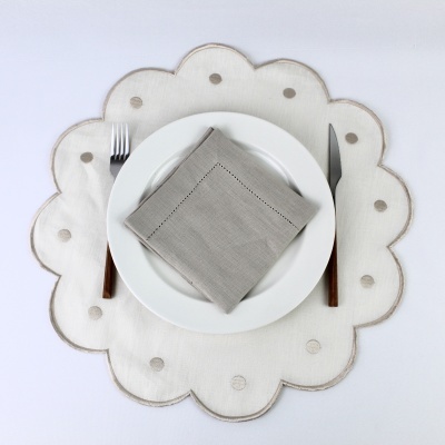 Conjunto de mesa com prato branco, guardanapo cinza, faca e garfo com cabos de madeira, sobre toalha branca com pontos cinzentos e bordas em forma de flor.