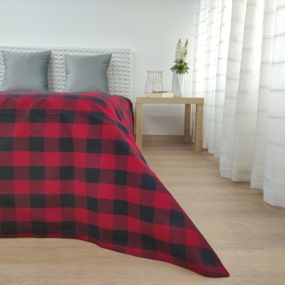 Cobertor xadrez vermelho e preto sobre cama branca com almofadas cinzentas e cortinas brancas ao lado